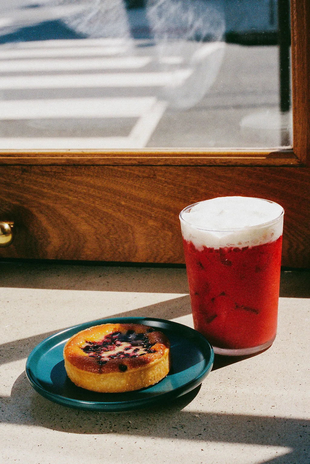 Red drink with foam and a pastry on a blue plate by a window
