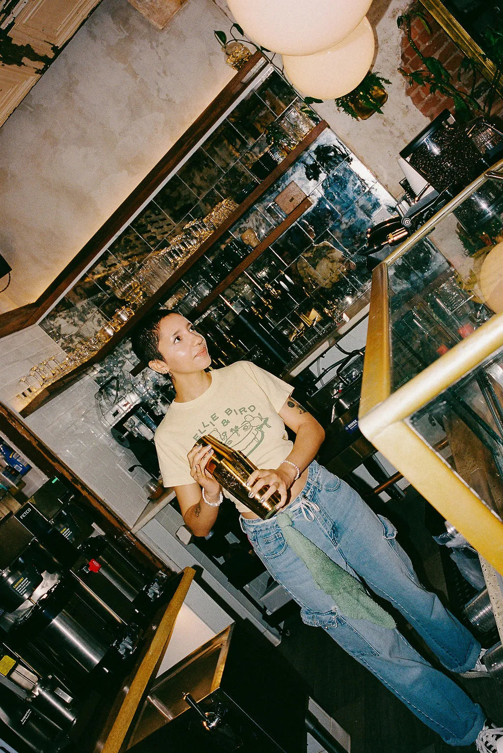 Person standing behind a bar counter with various bottles and glasses holding a gold shaker