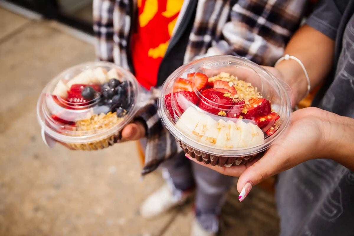 Two plastic containers of fruit bowls with berries and granola held by two people on a sidewalk.
