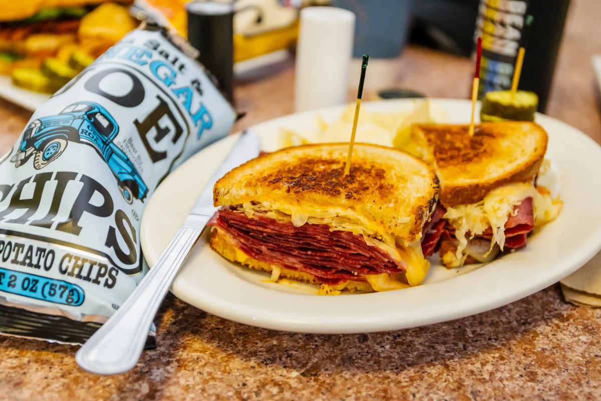 Grilled cheese sandwich with salami on a plate with a bag of chips and coffee in the background.