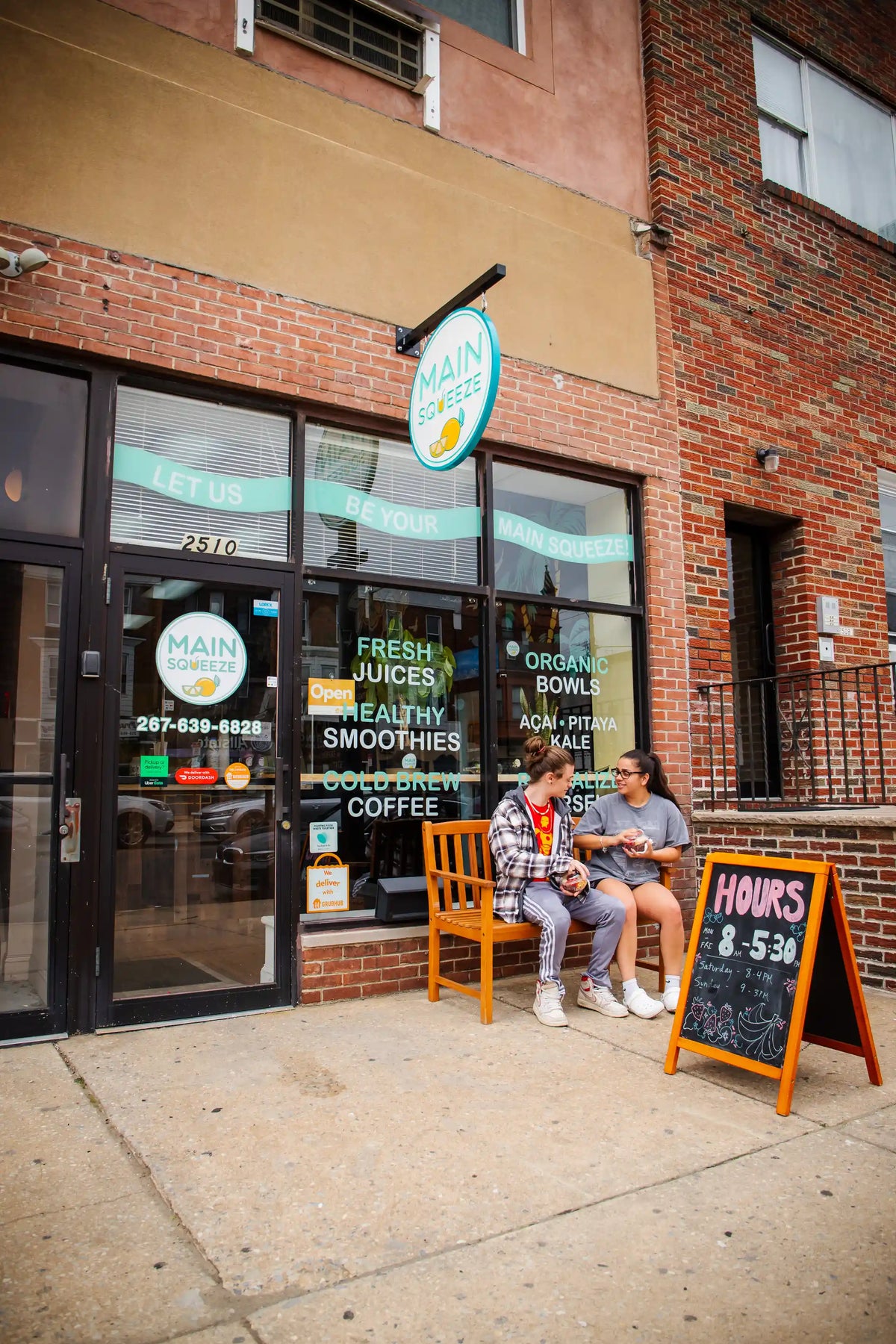 Two people sitting on a bench outside Main Squeeze storefront with a sign displaying hours.