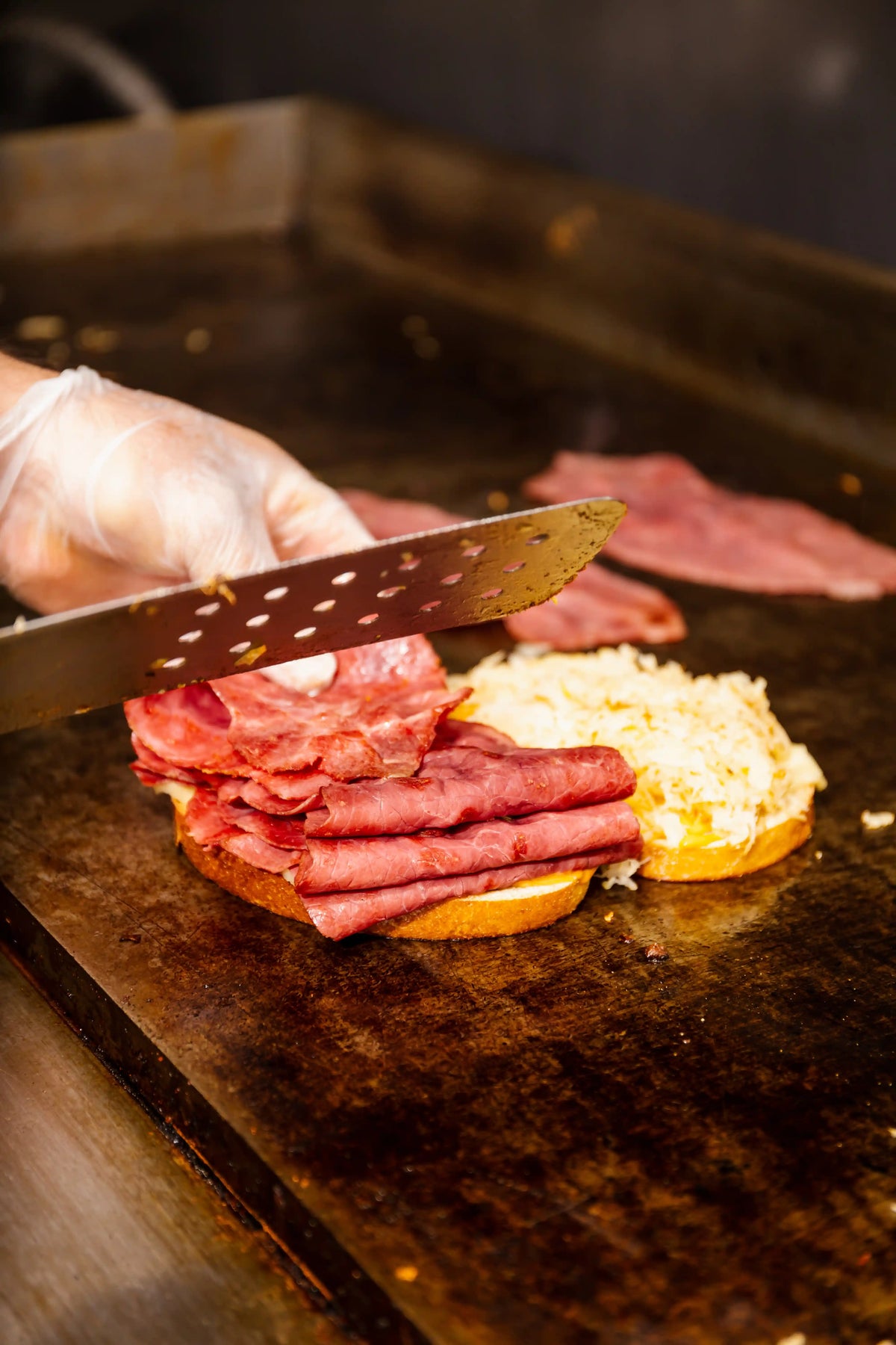 Person preparing a sandwich with sliced meats on a grill