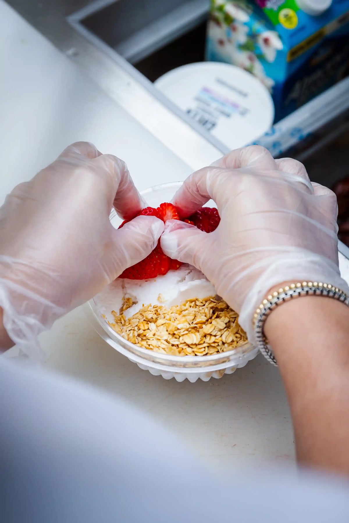 Person in gloves adding toppings to a fruit oat bowl on a kitchen counter.