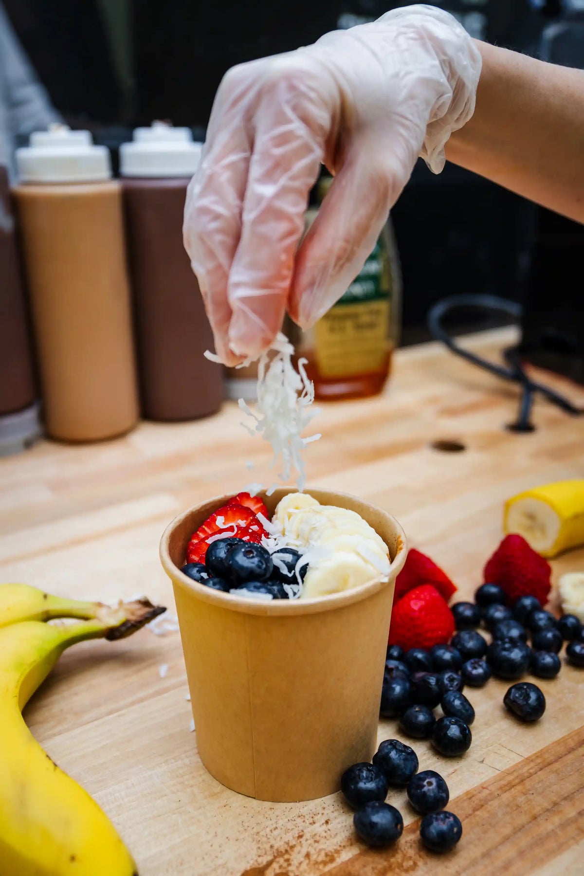 Hand with glove adding toppings to a cup of yogurt with fruit on a wooden surface