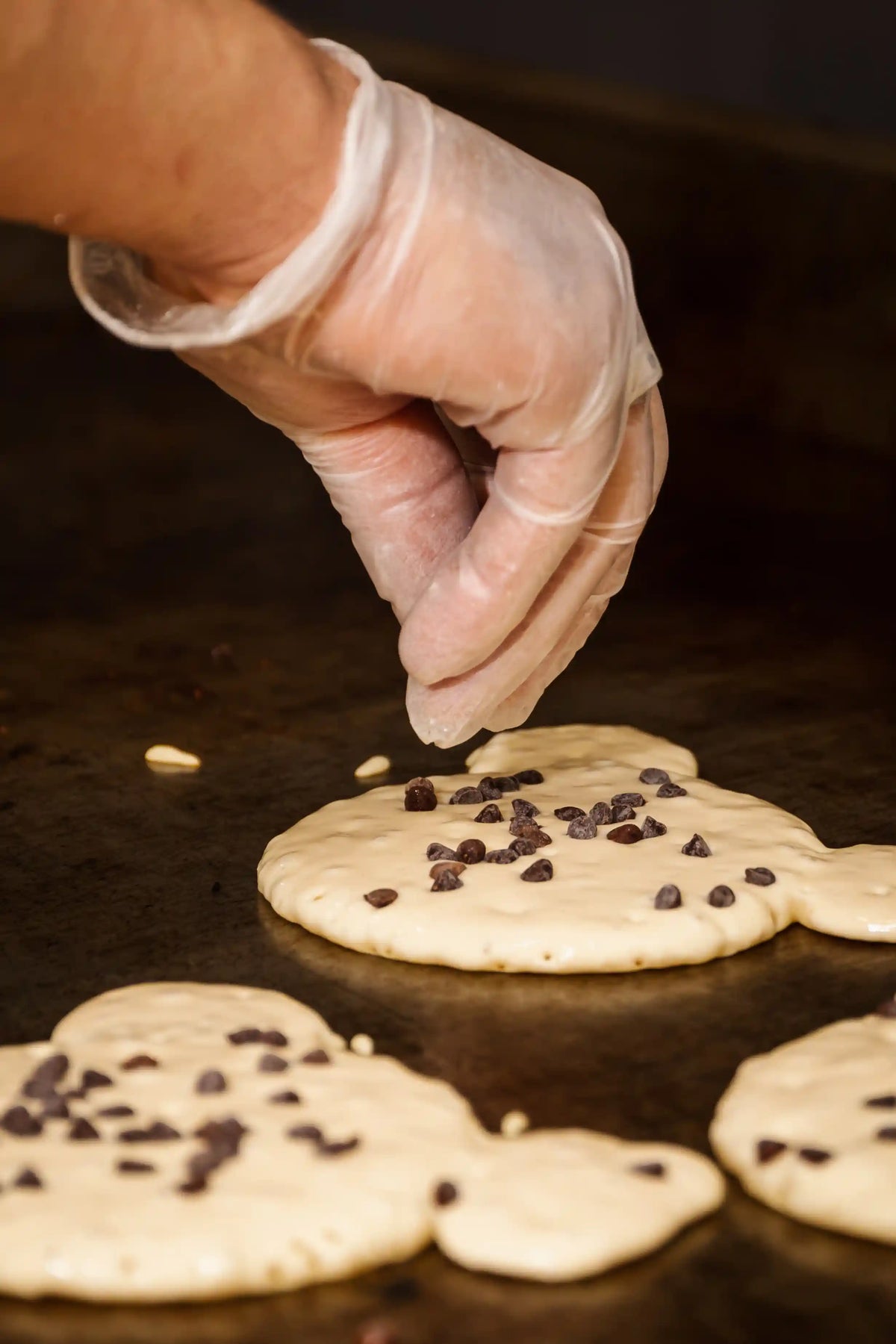 Hand sprinkling chocolate chips onto pancakes on a griddle.