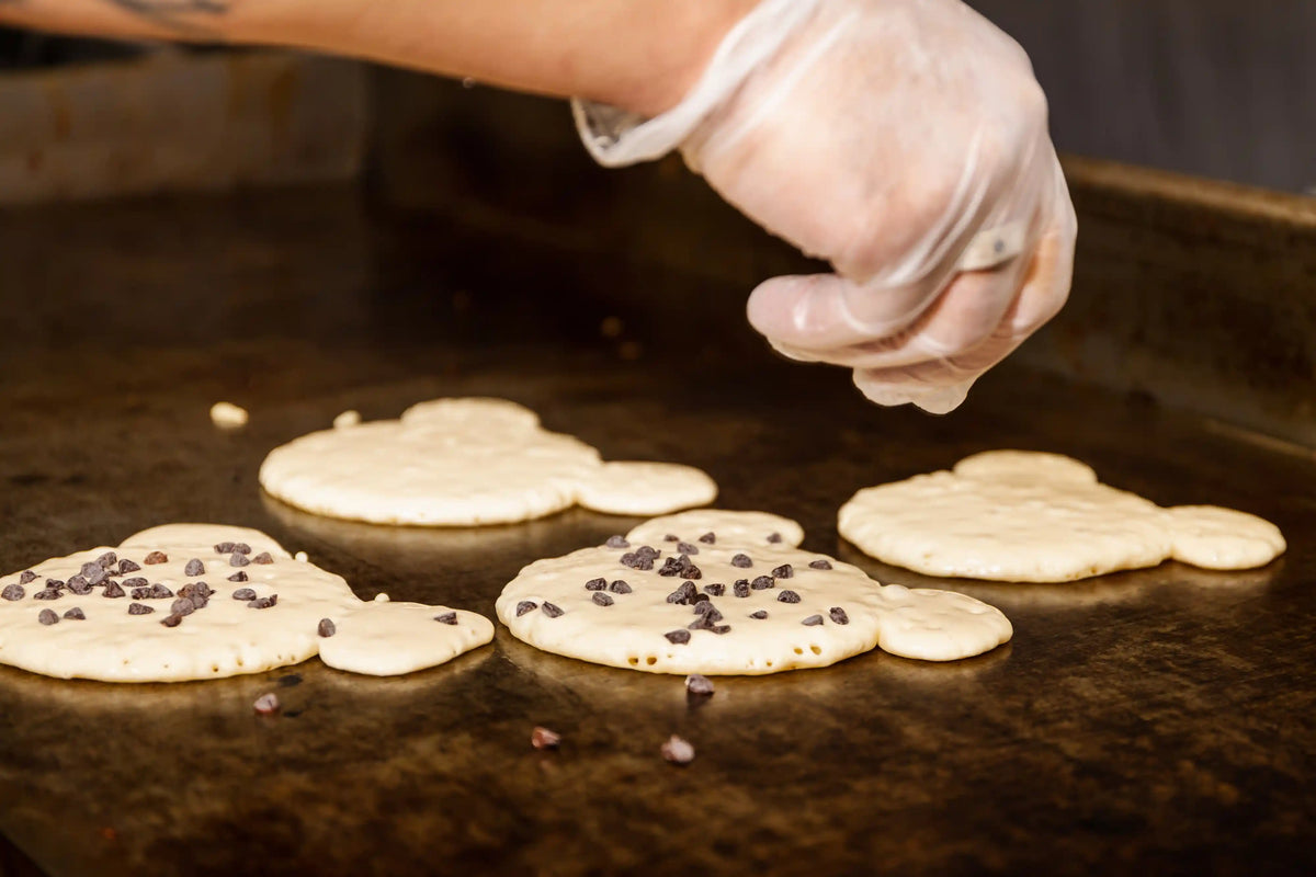 Person wearing gloves sprinkling chocolate chips onto pancakes on a griddle.