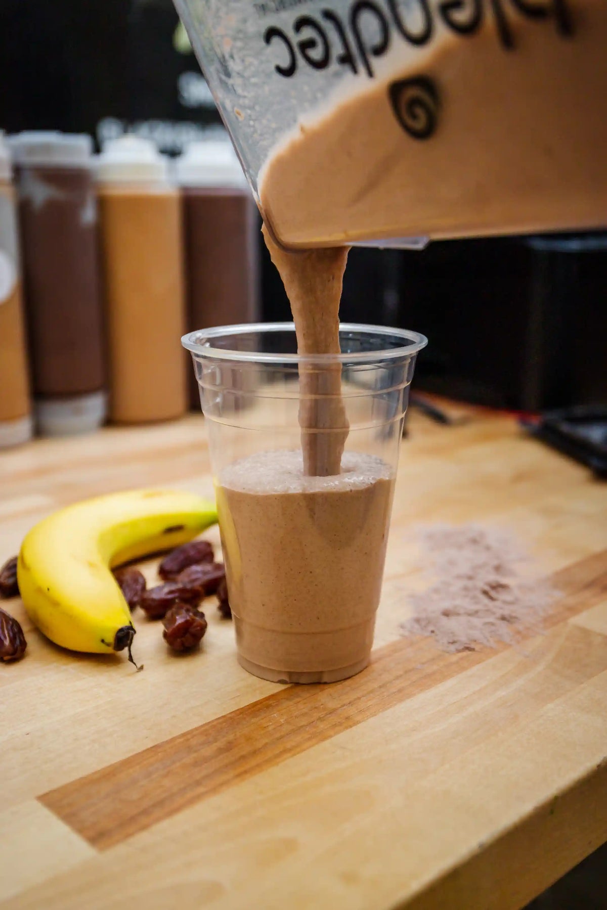 Smoothie being poured into a clear cup on a wooden surface with bananas and dates.