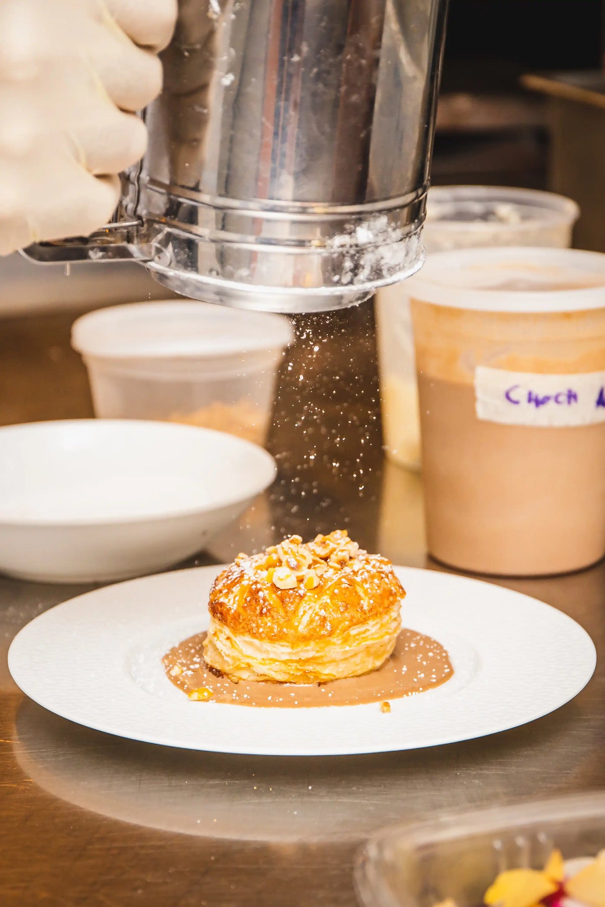 Person dusting powdered sugar over a dessert on a plate with a coffee cup in the background.