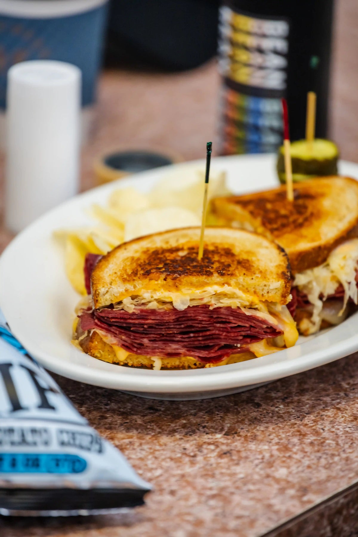 Grilled cheese sandwich with salami on a plate with chips and a drink in the background.