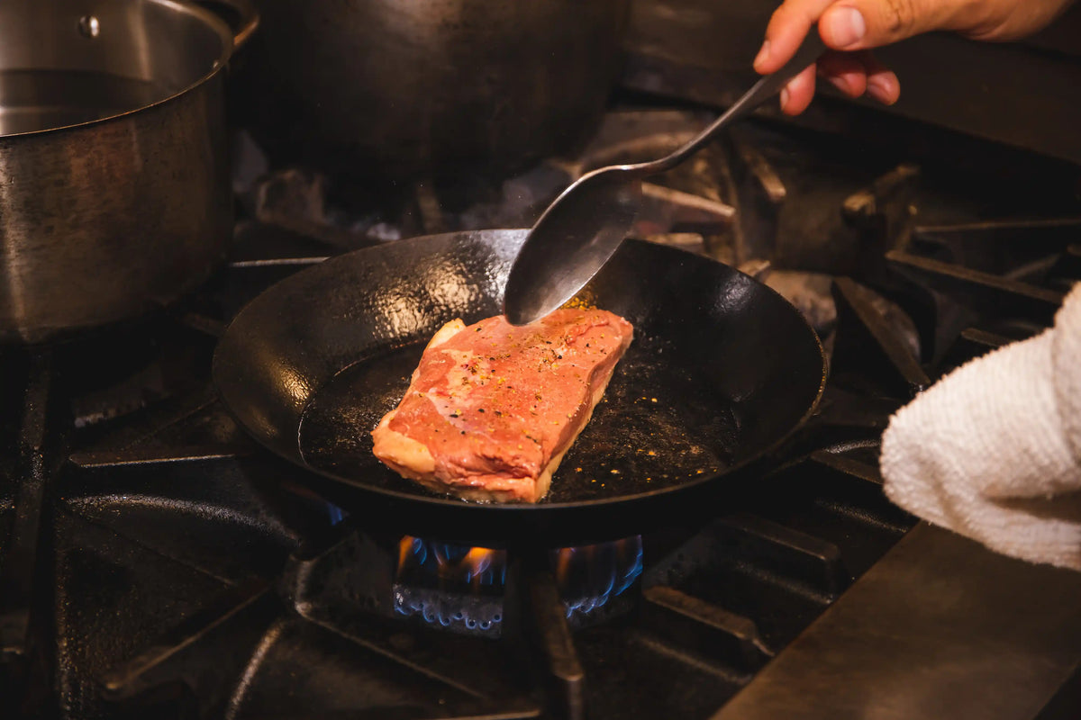 Person cooking a piece of meat in a frying pan on a professional stovetop.