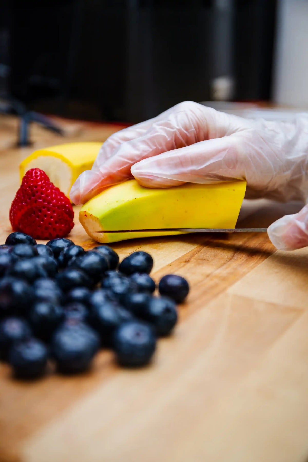 Person wearing gloves cutting a banana on a wooden cutting board with berries nearby.