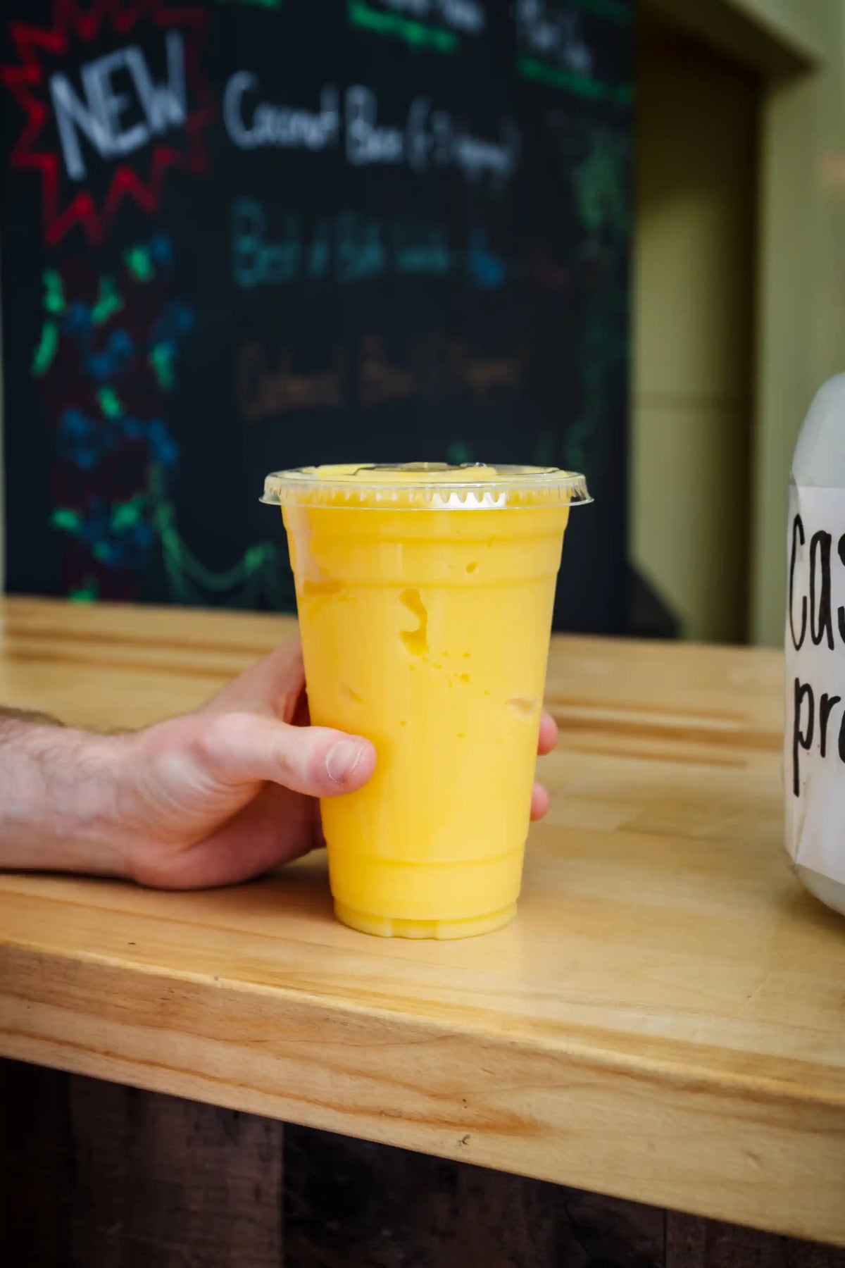 Hand holding a yellow smoothie drink in a clear plastic cup on a wooden counter with a chalkboard menu in the background.