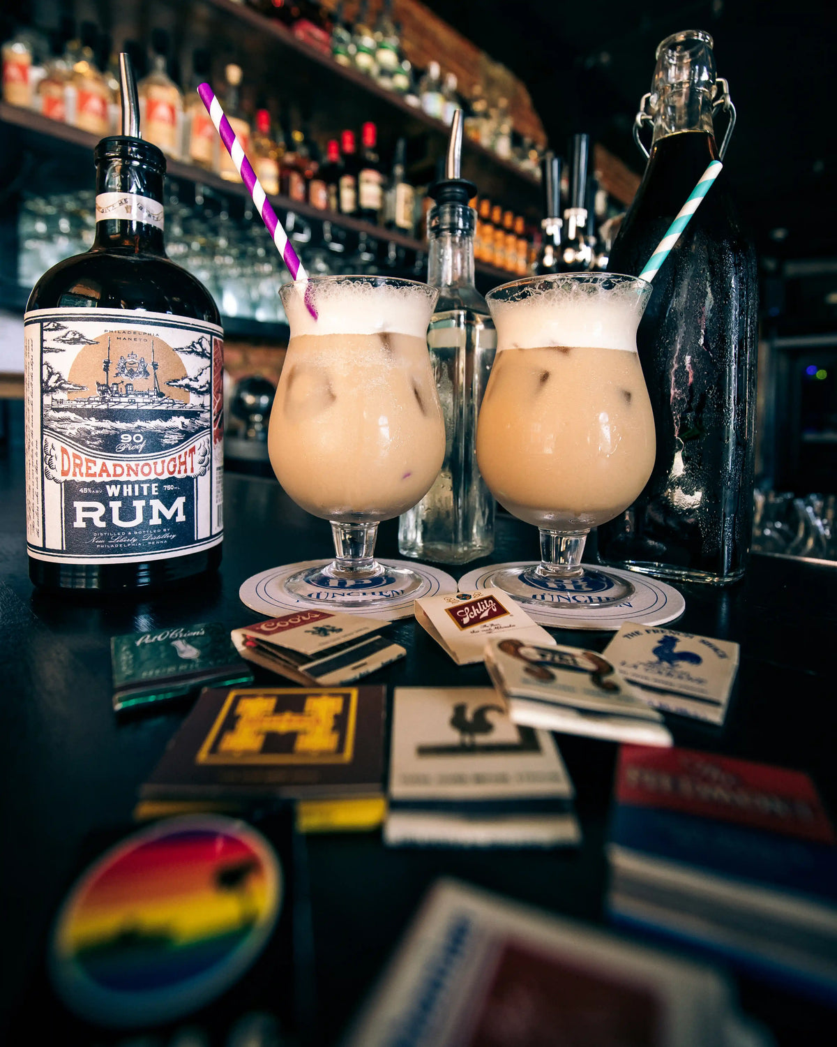 Two glasses of creamy cocktail with straws next to a bottle of Dreadnought Rum on a bar counter.